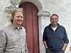 To menn står foran en hvit steinkirke med røde dører og ser mot kamera.