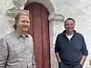 To menn står foran en hvit steinkirke med røde dører og ser mot kamera.